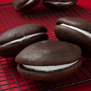 May include: Five chocolate sandwich cookies with white frosting on a black wire rack. The cookies are arranged in a semi-circle on the rack.