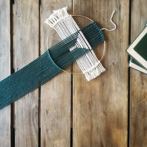Puede incluir: Un tapiz de macramé verde y blanco con un marco circular de madera. El macramé está tejido en un patrón geométrico.