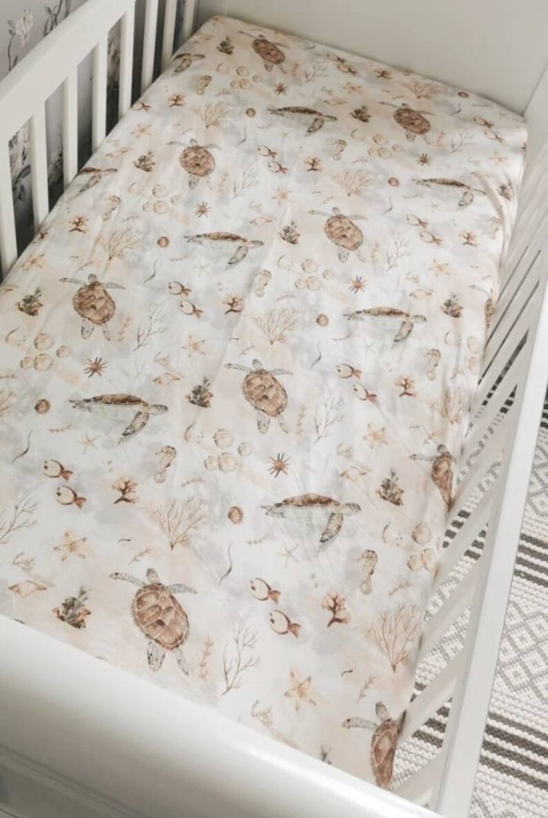 May include: A white crib sheet with a repeating pattern of sea turtles, fish, seahorses, and coral in shades of brown and beige. The sheet is made of soft fabric and is designed to fit a standard crib.