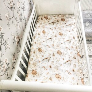 May include: A white crib with a patterned sheet featuring sea turtles, fish, and coral in shades of brown and beige. The crib is set against a floral wallpaper background.