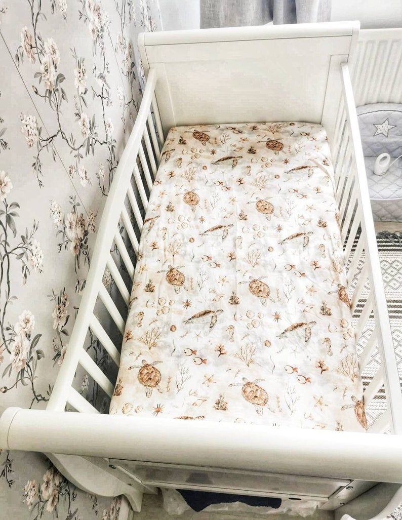 May include: A white crib with a patterned sheet featuring sea turtles, fish, and coral in shades of brown and beige. The crib is set against a floral wallpaper background.