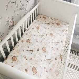 May include: A white crib with a fitted sheet featuring a sea turtle pattern. The sheet has a cream background with brown sea turtles, fish, and coral. The crib is set against a floral wallpaper.