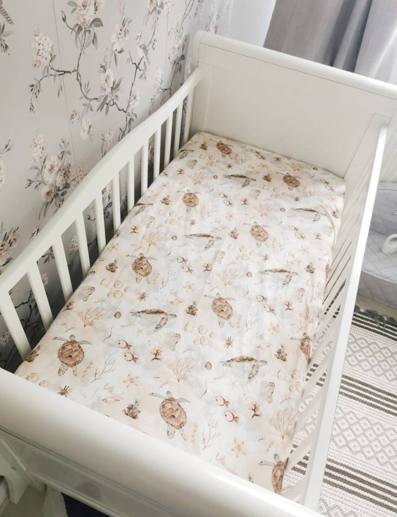 May include: A white crib with a fitted sheet featuring a sea turtle pattern. The sheet has a cream background with brown sea turtles, fish, and coral. The crib is set against a floral wallpaper.