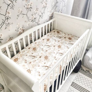 May include: A white cot with a patterned sheet featuring sea turtles, fish, and coral. The cot is set against a floral wallpaper background. The cot has a slatted design and a curved headboard.
