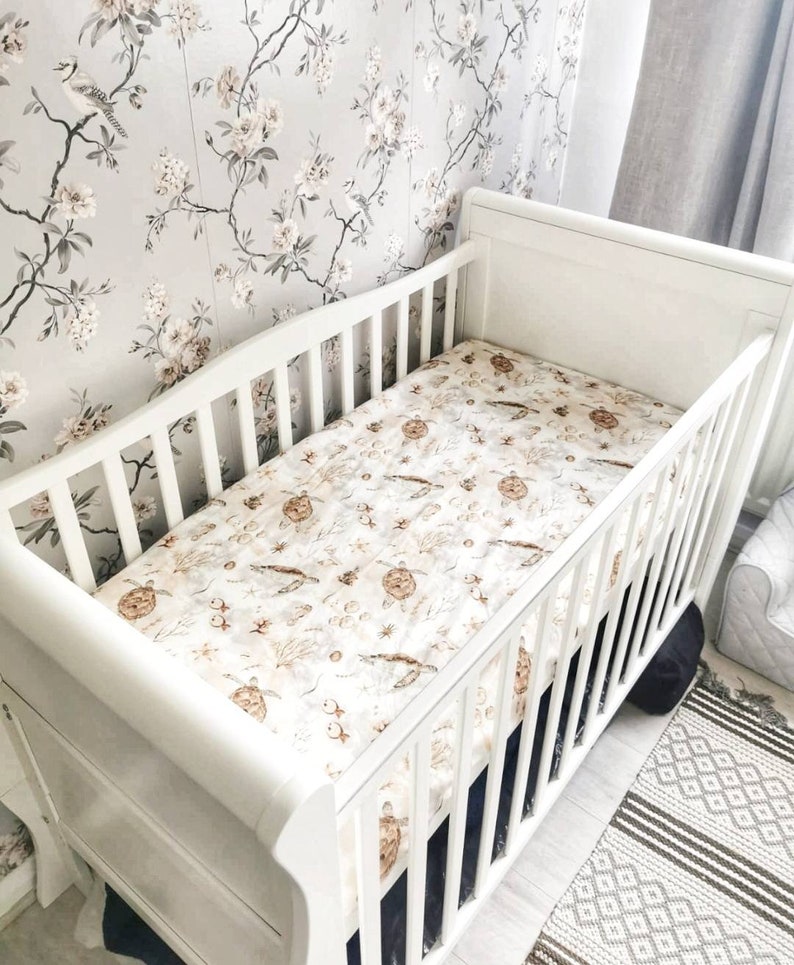 May include: A white crib with a patterned sheet featuring sea turtles, fish, and coral. The crib is set against a floral wallpaper background. The crib has a slatted design and a curved headboard.