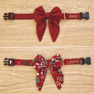 Pode incluir: Duas coleiras para cães com laços. Uma é bordô lisa, a outra é bordô com um padrão festivo de folhas verdes e bagas vermelhas. Ambas têm fivelas de plástico preto e uma pequena etiqueta com texto.