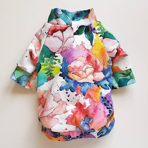 May include: A white dog sweater with a colorful floral print. The print features watercolor-style flowers and leaves in shades of pink, blue, green, and yellow.