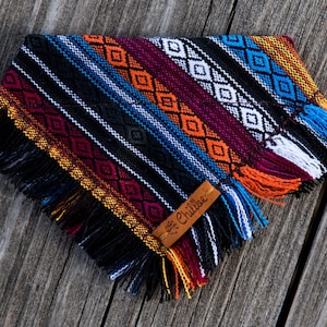 May include: A colourful dog bandana with a black background and a geometric pattern in shades of red, orange, yellow, blue, and white. The bandana has fringe around the edges and a brown leather tag with the text "Chillbee" on it.