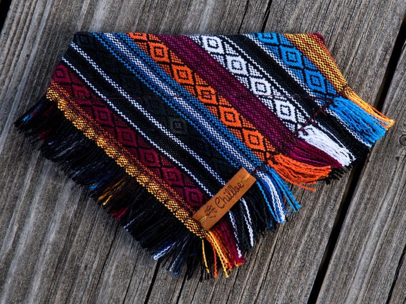 May include: A colourful dog bandana with a black background and a geometric pattern in shades of red, orange, yellow, blue, and white. The bandana has fringe around the edges and a brown leather tag with the text "Chillbee" on it.