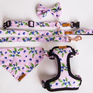 Op de afbeelding: Een bijpassende set accessoires voor huisdieren met een lichtroze achtergrond en een blauwe bessen- en bladerenpatroon. De set bevat een vlinderdas, halsband, riem, bandana en harnas. Het harnas heeft zwarte randen en hardware.