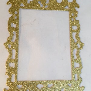 May include: A gold glitter picture frame with an ornate, decorative design. The frame is rectangular with a detailed, textured border. The frame is empty, with a white background visible through the opening.