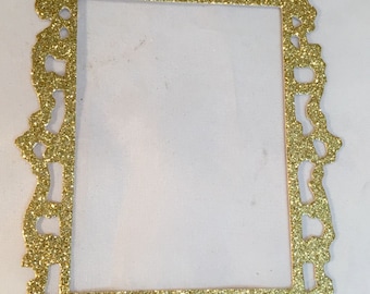 May include: A gold glitter picture frame with an ornate, decorative design. The frame is rectangular with a detailed, textured border. The frame is empty, with a white background visible through the opening.