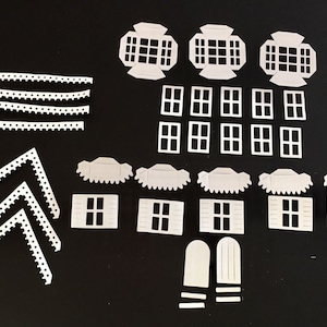 May include: A collection of white paper cutouts for a miniature house project. The set includes roof pieces, windows, decorative trim, and house walls. The pieces are arranged on a black surface, ready for assembly.