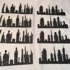 May include: Eight black paper cutouts of city skylines. Each cutout features a unique arrangement of buildings, towers, and architectural details. The silhouettes are displayed against a white background.