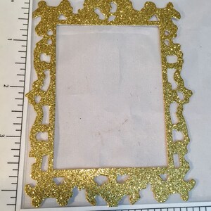 May include: A gold glitter picture frame with ornate detailing. The frame is rectangular with a decorative top and bottom edge. The frame measures approximately 3 inches in height.