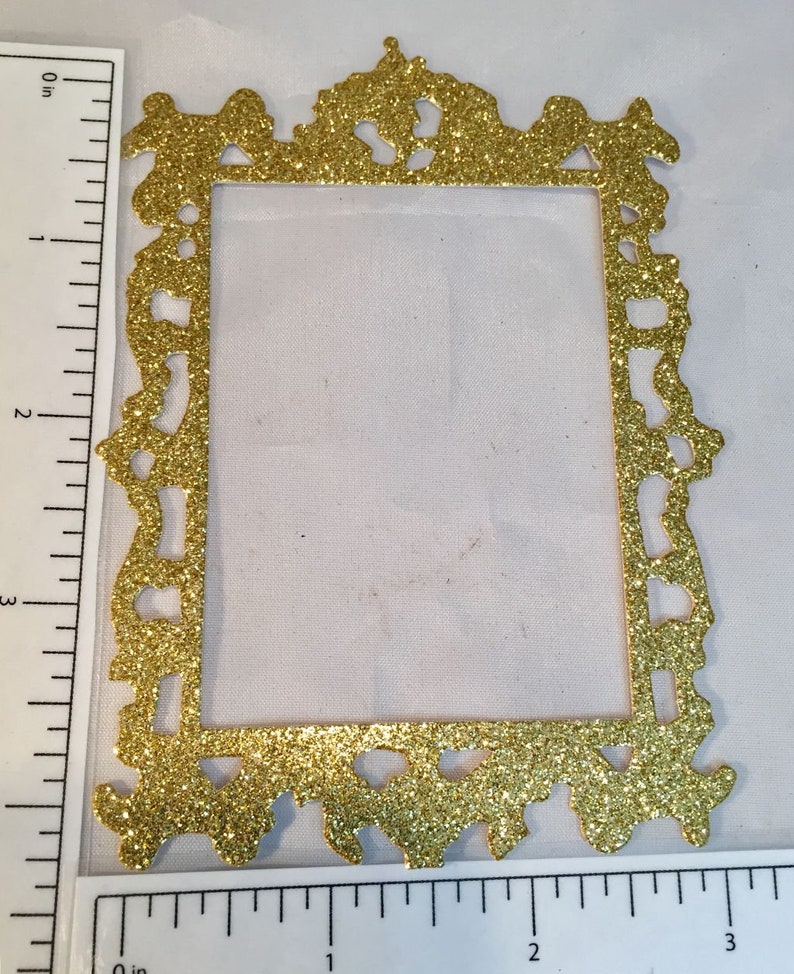May include: A gold glitter picture frame with ornate detailing. The frame is rectangular with a decorative top and bottom edge. The frame measures approximately 3 inches in height.