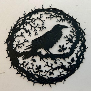 May include: A black raven silhouette perched on a branch within a circular frame of thorny branches. The raven is detailed with a beak and eye, set against a white background. The artwork has a gothic or nature-inspired theme.