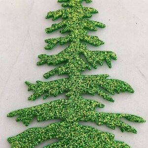 May include: A glittery green Christmas tree decoration. The tree is cut out in the shape of a fir tree with detailed branches. The glitter is a mix of green and gold, giving it a festive look. The tree is set against a white background.