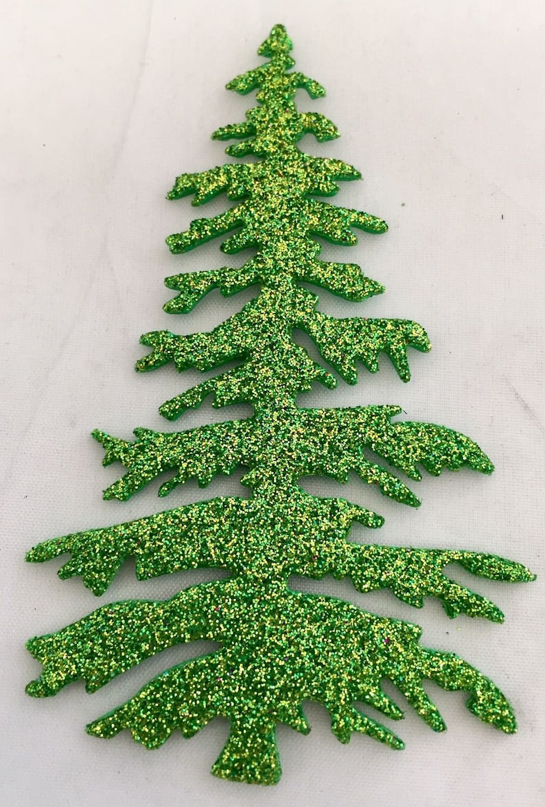 May include: A glittery green Christmas tree decoration. The tree is cut out in the shape of a fir tree with detailed branches. The glitter is a mix of green and gold, giving it a festive look. The tree is set against a white background.