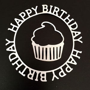 May include: A white, die-cut paper design with the words "HAPPY BIRTHDAY" encircling a cupcake silhouette. The cupcake has a detailed paper cut design. The background is solid black, providing contrast to the white design.