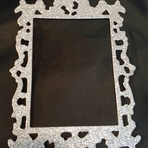 May include: A silver glitter picture frame with an ornate, decorative design. The frame has a rectangular opening and a detailed, textured border. The frame is made of a shiny, glittery material.