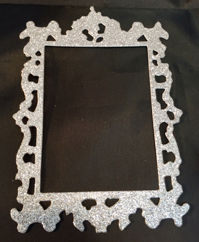 May include: A silver glitter picture frame with an ornate, decorative design. The frame has a rectangular opening and a detailed, textured border. The frame is made of a shiny, glittery material.