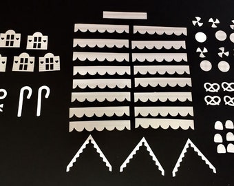 May include: A collection of white, die-cut pieces for a gingerbread house, including roof sections, walls, windows, and decorative elements like pretzels, bows, and candy canes. The pieces are arranged on a black surface.