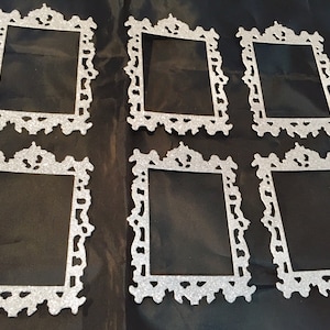 May include: Six silver glitter picture frames with ornate, decorative edges are arranged on a black surface. The frames have a rectangular opening and a detailed, vintage-inspired design, suitable for crafts or home decor.