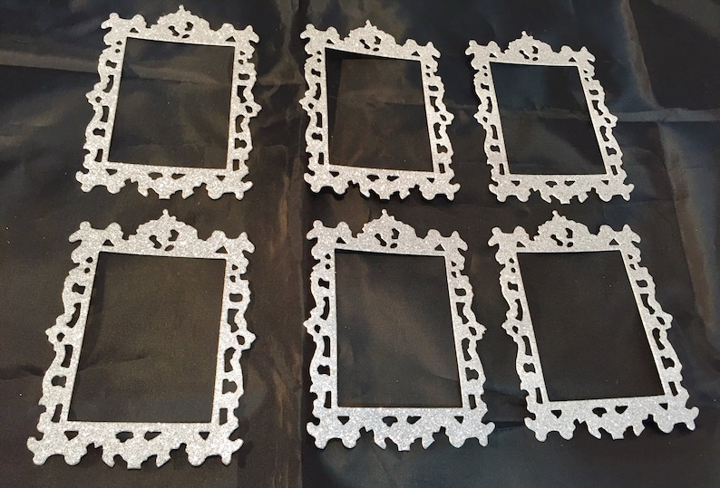 May include: Six silver glitter picture frames with ornate, decorative edges are arranged on a black surface. The frames have a rectangular opening and a detailed, vintage-inspired design, suitable for crafts or home decor.