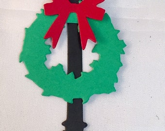 May include: A black silhouette of a lamppost with a green wreath and a red bow. The lamppost has a decorative top and a flared base. The wreath is circular and has a scalloped edge.