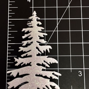 May include: A glittery white Christmas tree-shaped craft embellishment. The tree is approximately 3.5 inches tall and has a textured, sparkly surface. The tree is set against a black grid background, with a ruler for scale.