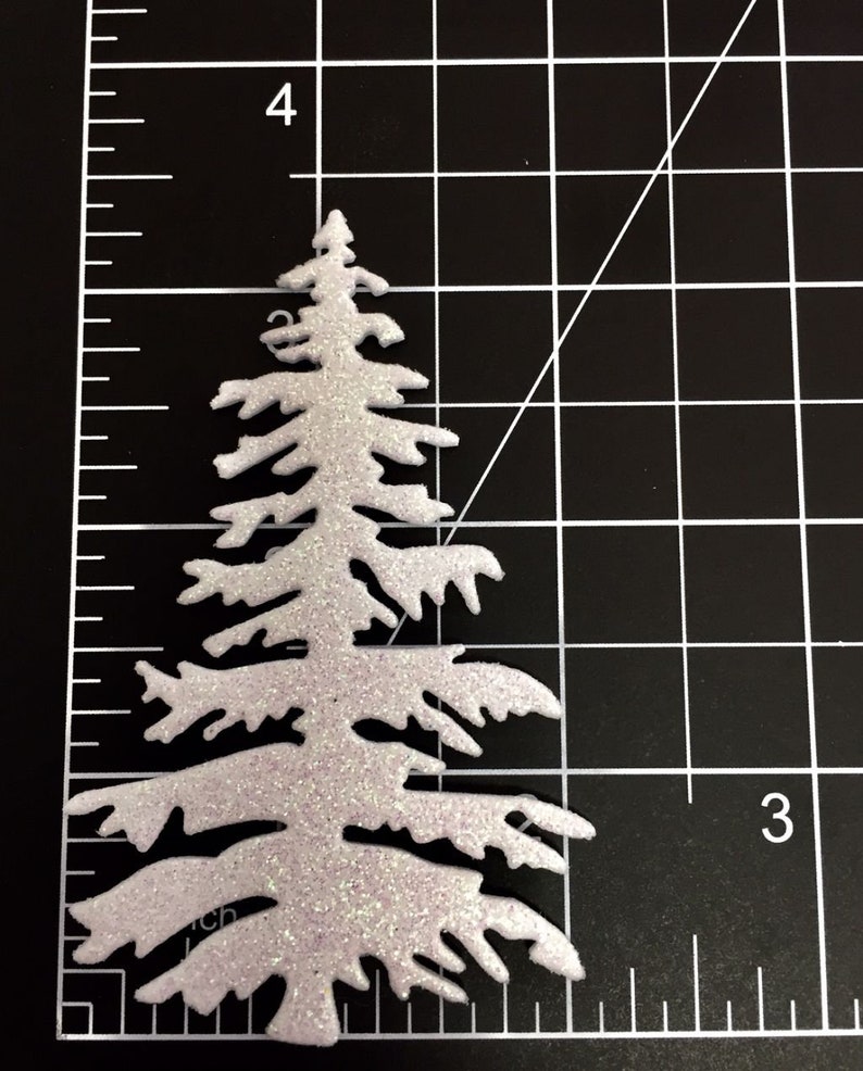 May include: A glittery white Christmas tree-shaped craft embellishment. The tree is approximately 3.5 inches tall and has a textured, sparkly surface. The tree is set against a black grid background, with a ruler for scale.