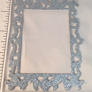 May include: A silver glitter-covered decorative frame with an ornate, vintage design. The frame is rectangular with a central opening and decorative flourishes along the edges. The frame is approximately 3 inches tall.