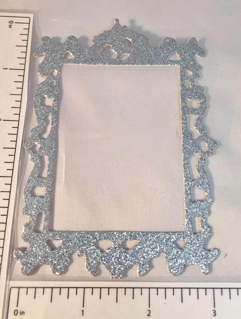May include: A silver glitter-covered decorative frame with an ornate, vintage design. The frame is rectangular with a central opening and decorative flourishes along the edges. The frame is approximately 3 inches tall.