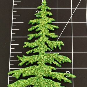 May include: A glittery green Christmas tree-shaped embellishment. The tree is cut out with detailed branches and a textured, sparkling surface. The tree is approximately 3 inches tall and is set against a black background with a ruler.