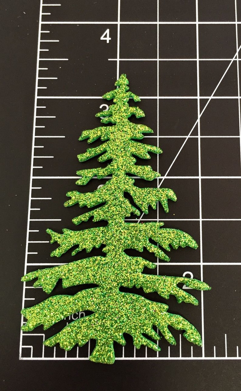 May include: A glittery green Christmas tree-shaped embellishment. The tree is cut out with detailed branches and a textured, sparkling surface. The tree is approximately 3 inches tall and is set against a black background with a ruler.