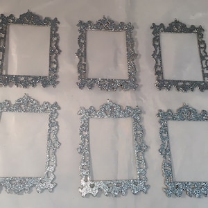 May include: Six silver glitter picture frames with ornate, decorative edges. The frames are rectangular with a clear center, perfect for displaying photos or artwork. The frames are arranged on a white surface.