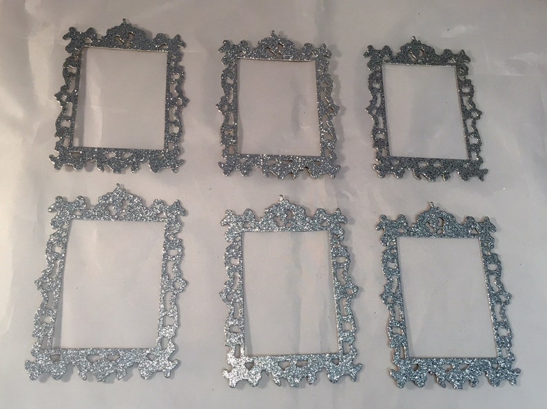 May include: Six silver glitter picture frames with ornate, decorative edges. The frames are rectangular with a clear center, perfect for displaying photos or artwork. The frames are arranged on a white surface.