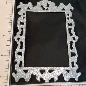 May include: A silver glitter picture frame with an ornate, decorative design. The frame is rectangular with a cutout center and measures approximately 4 inches in height.