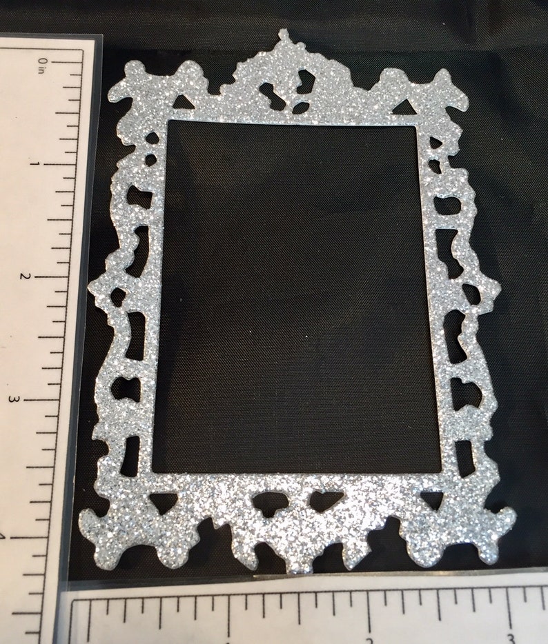 May include: A silver glitter picture frame with an ornate, decorative design. The frame is rectangular with a cutout center and measures approximately 4 inches in height.