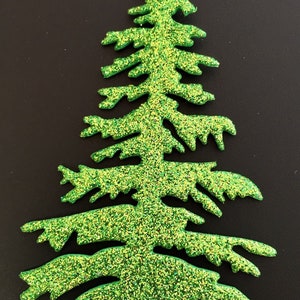 May include: A glittery green Christmas tree-shaped decoration. The tree has a classic evergreen shape with detailed branches and is covered in sparkling green and yellow glitter. The background is a solid black.