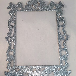 May include: A silver glitter picture frame with an ornate, decorative design. The frame is rectangular with a detailed border and a blank center. The glitter adds a sparkling effect to the frame.