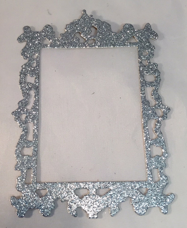 May include: A silver glitter picture frame with an ornate, decorative design. The frame is rectangular with a detailed border and a blank center. The glitter adds a sparkling effect to the frame.