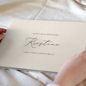 May include: A white linen guest book with the text "Happy 60th Birthday Kristine Love from your girls x" printed in black.