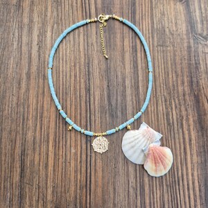 May include: A light blue beaded necklace featuring small gold-toned spacers and a central gold-toned coin pendant. The necklace includes a gold-toned lobster clasp and an adjustable chain extender, accompanied by two white and pink seashells.