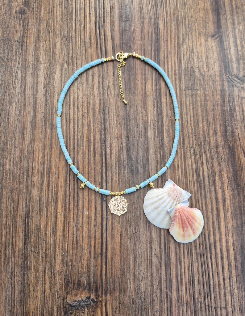 May include: A light blue beaded necklace featuring small gold-toned spacers and a central gold-toned coin pendant. The necklace includes a gold-toned lobster clasp and an adjustable chain extender, accompanied by two white and pink seashells.