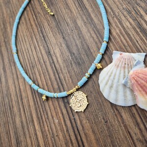 May include: A light blue beaded necklace featuring small, flat, circular turquoise-colored beads interspersed with tiny gold-toned metallic spacers. A circular gold-toned coin pendant hangs from the center, accompanied by two small gold-toned charms.