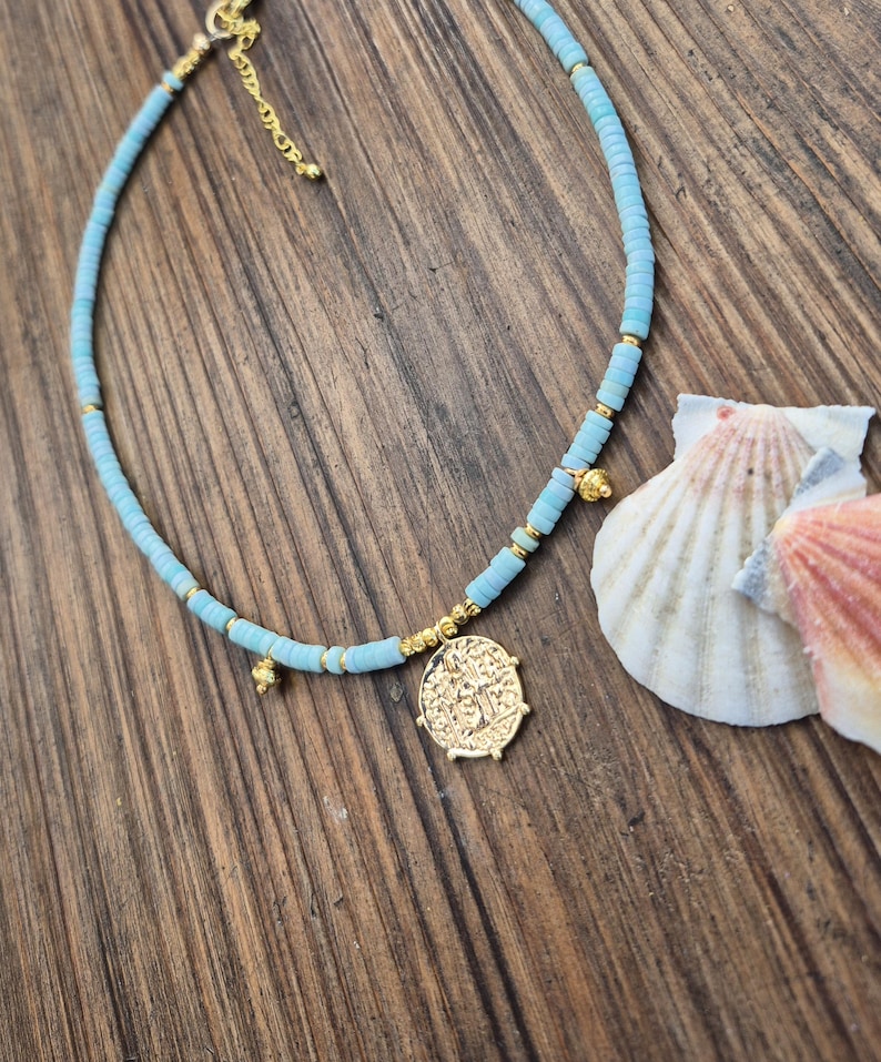 May include: A light blue beaded necklace featuring small, flat, circular turquoise-colored beads interspersed with tiny gold-toned metallic spacers. A circular gold-toned coin pendant hangs from the center, accompanied by two small gold-toned charms.