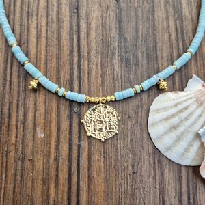 May include: A necklace featuring light blue disc-shaped beads interspersed with small gold-toned spherical beads. The necklace includes two small gold-toned charms and a central circular gold-toned pendant with an embossed design.