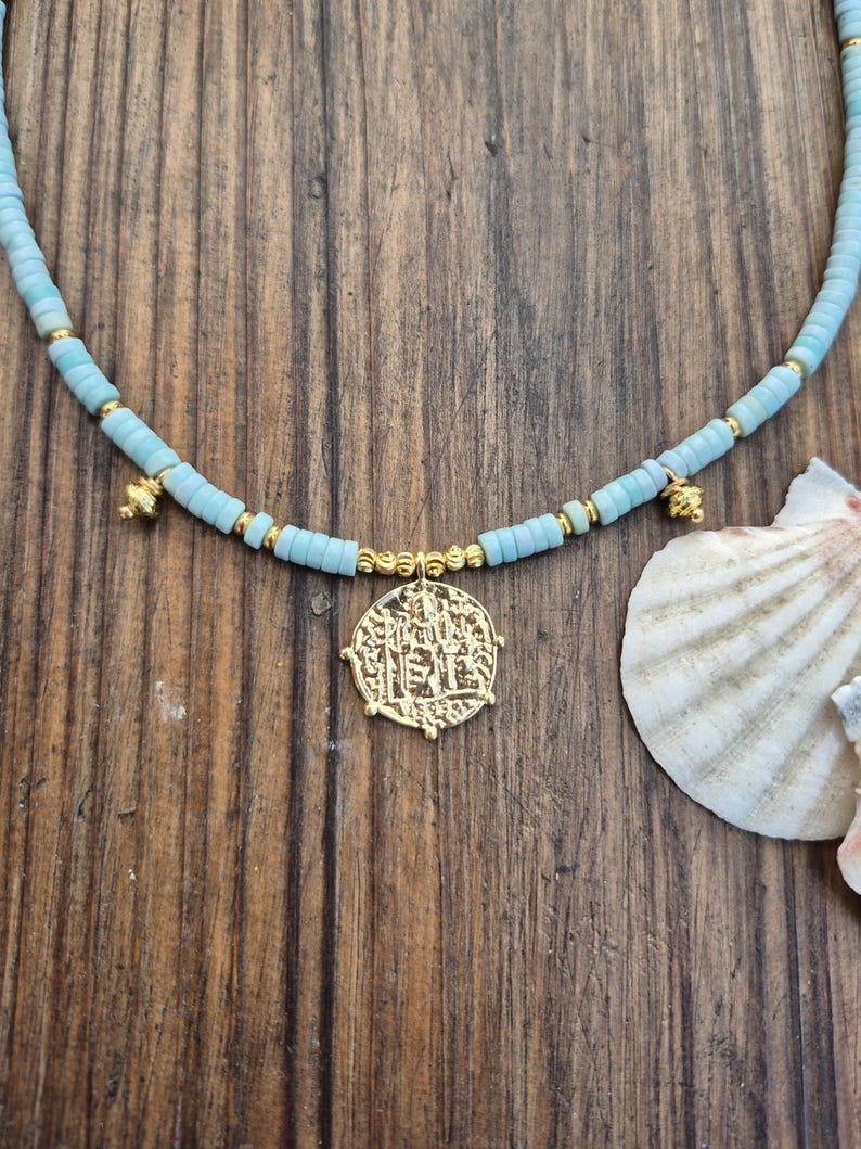 May include: A necklace featuring light blue disc-shaped beads interspersed with small gold-toned spherical beads. The necklace includes two small gold-toned charms and a central circular gold-toned pendant with an embossed design.
